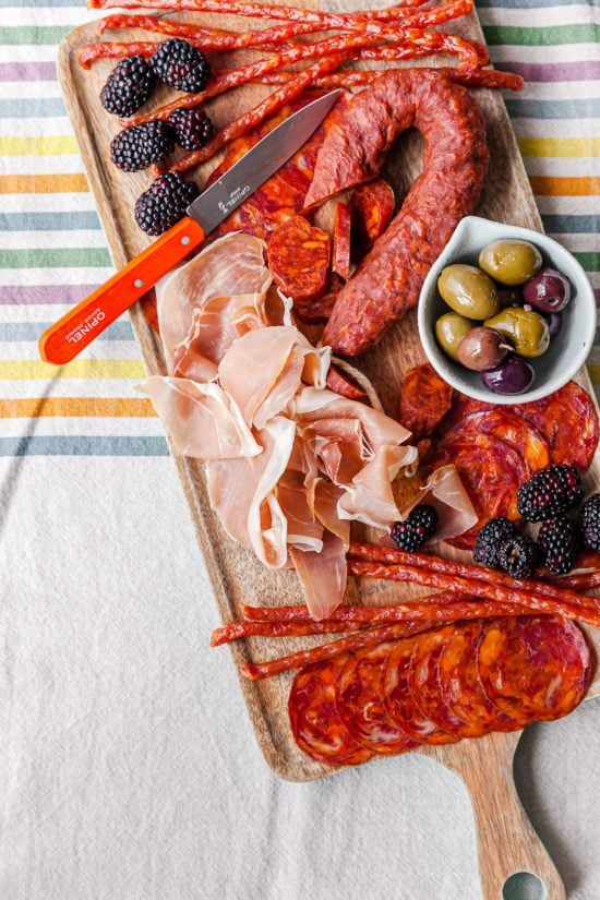 Knife on a wooden board with meats, olives, and fruit.