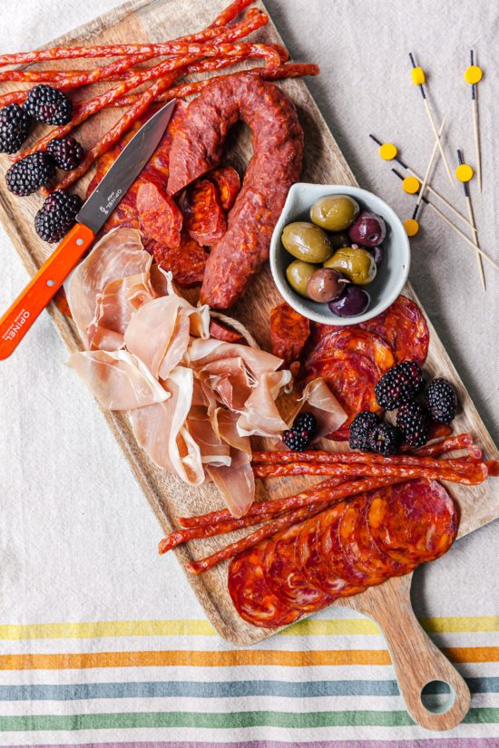 Meats, fruits, and olives on a wooden board.