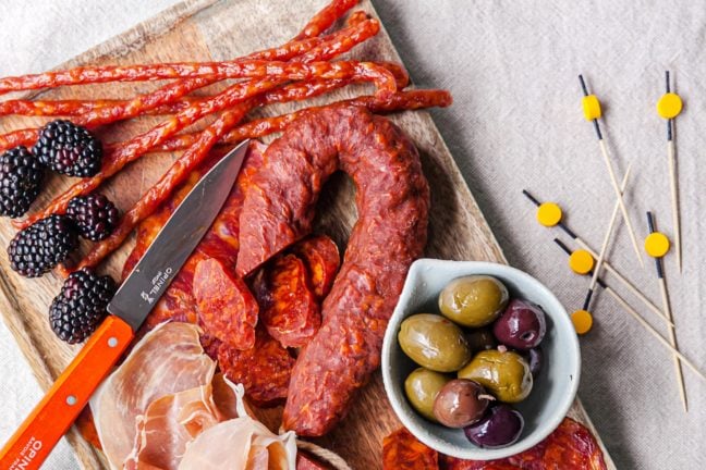 Various forms of meat on a wooden board with fruit and a bowl of olives.