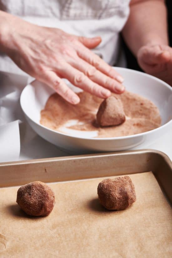 Woman rolling cookie dough in cinnamon sugar and placing them on a lined baking sheet.