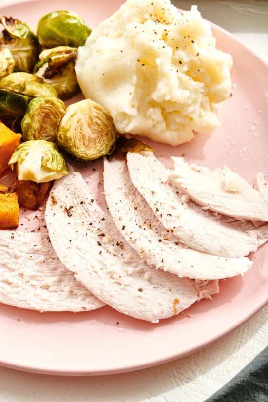 Slices of Turkey Breast on a plate with sweet potato, mashed potatoes, and brussels sprouts.