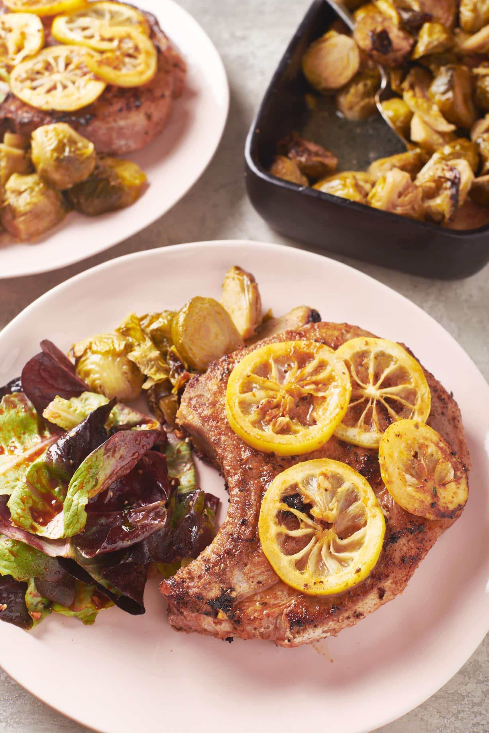Spanish Pork Chop topped with lemon slices on a plate with brussels sprouts and salad.
