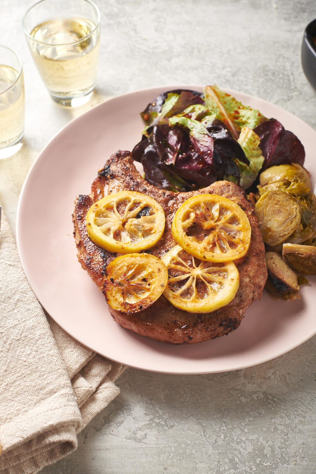 Lemon slices atop a Spanish Pork Chop on a plate with salad and brussels sprouts.