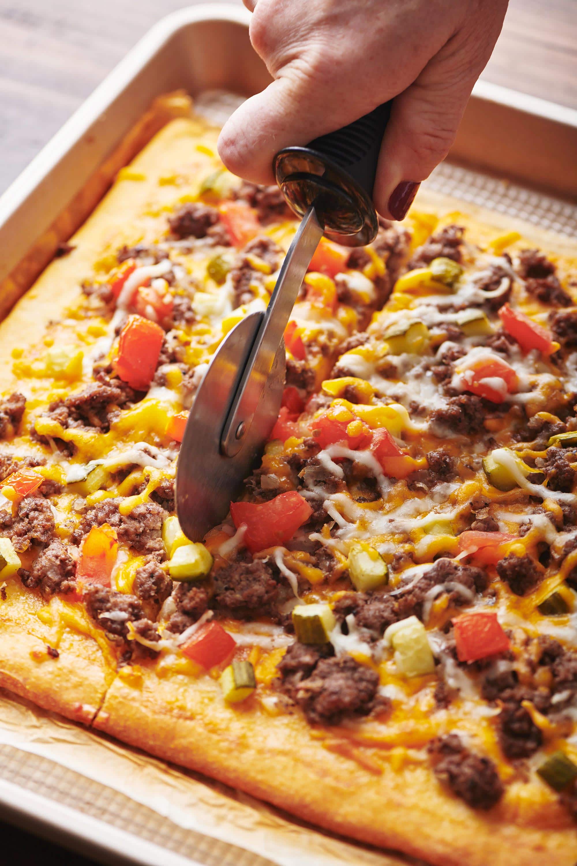 Woman using a pizza cutter to slice a Cheeseburger Pizza.