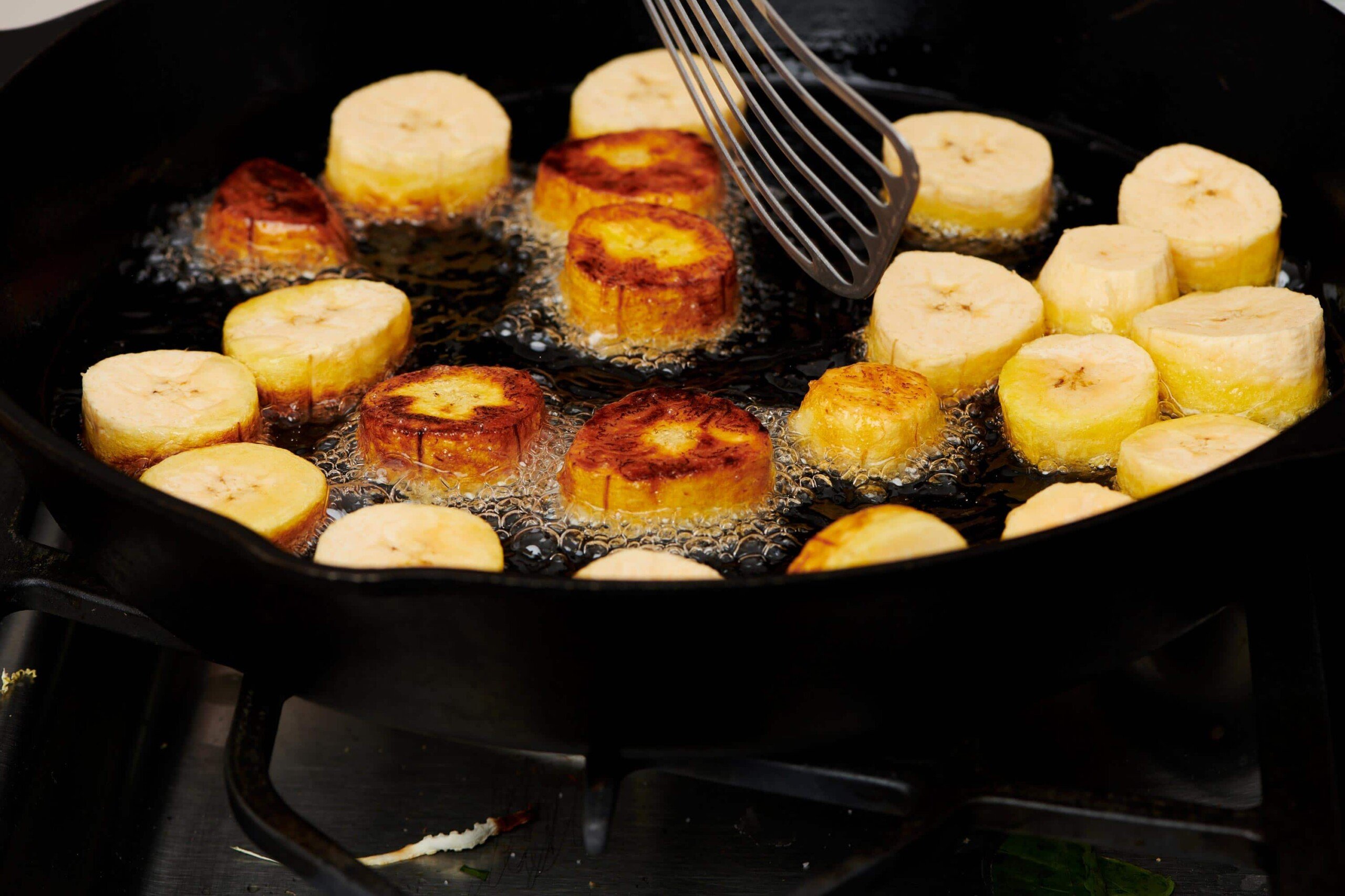 Metal spatula flipping plantain slices in a skillet.