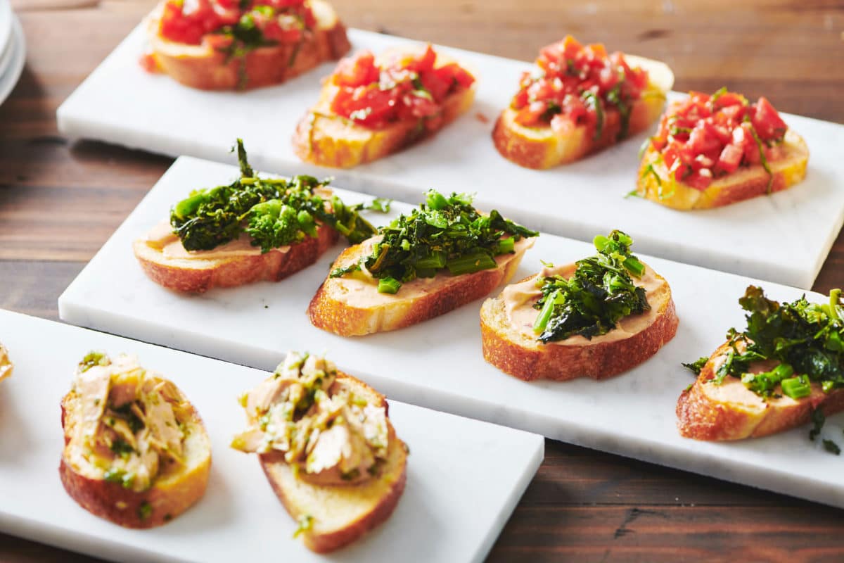 Broccoli Rabe Crostini with Chipotle Sauce on a long, white plate between plates of other crostini.