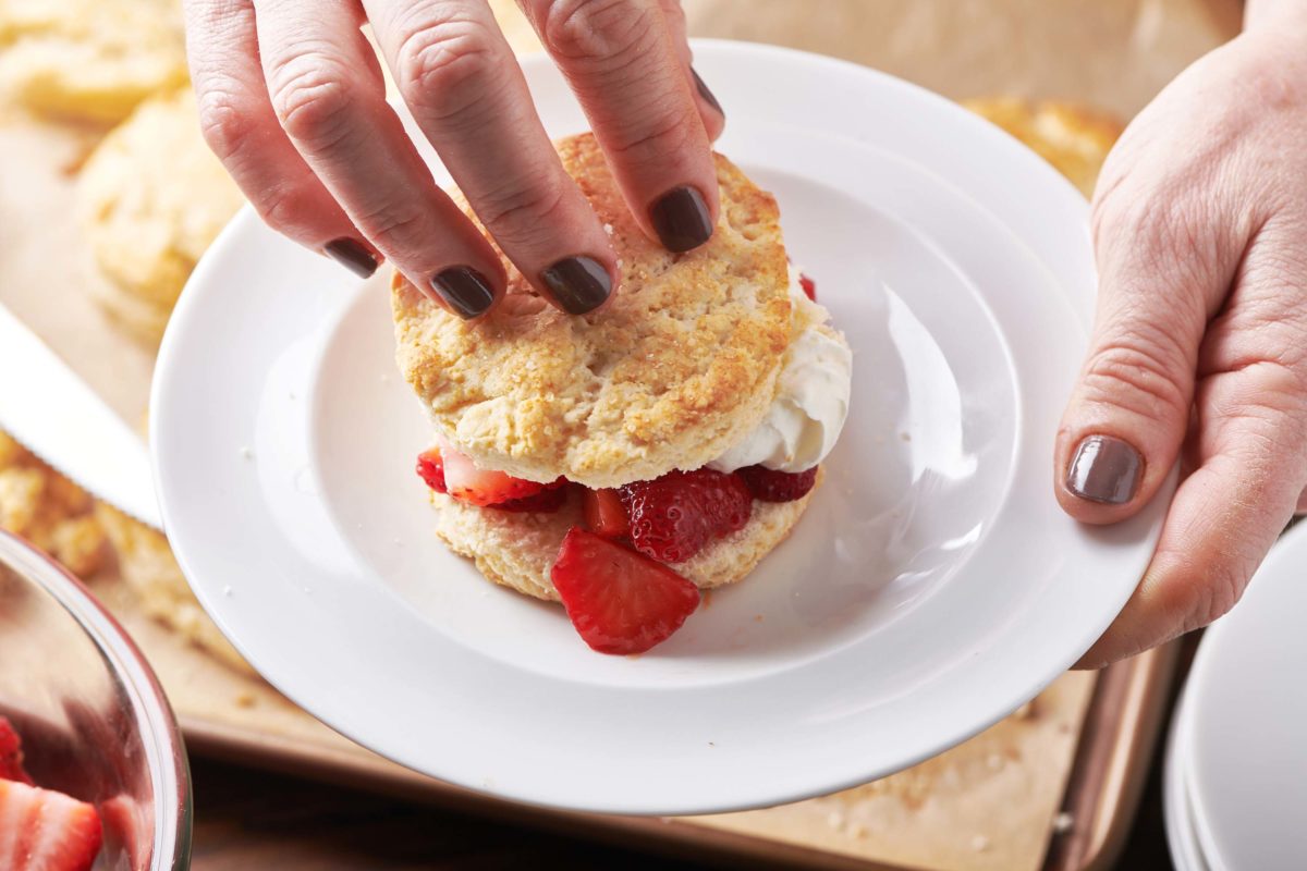 Woman adding a biscuit on top of layers of whipped cream, strawberries, and biscuit.