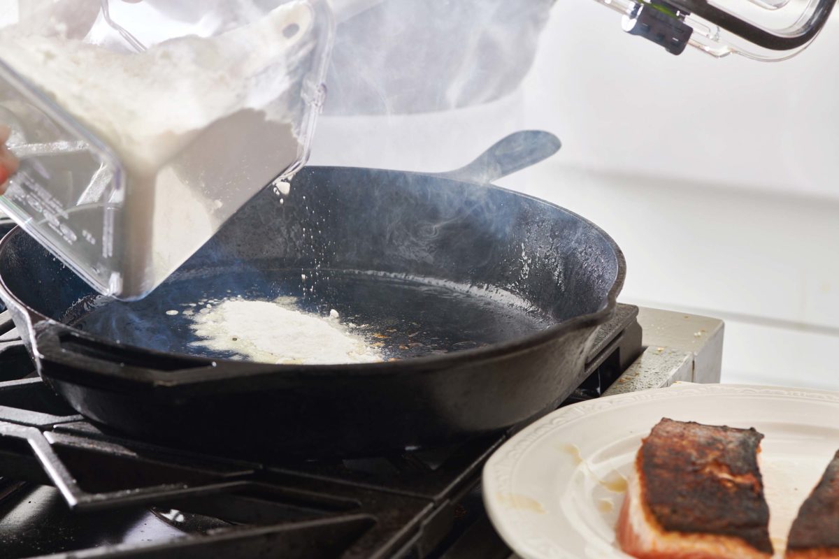 Flour pouring into a skillet next to a plate of salmon.