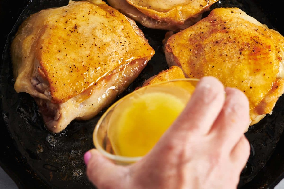 Woman pouring melted butter onto chicken thighs in a skillet.