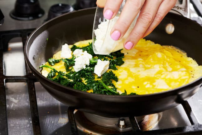 Woman pouring cheese onto an omelet in a skillet.