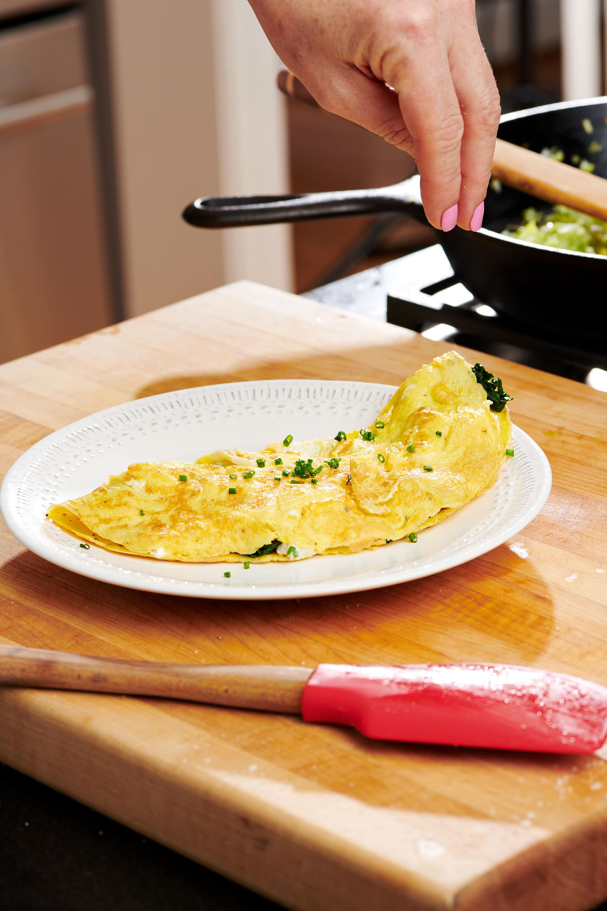 Woman sprinkling herbs on a Omelet on a white plate on a wooden board.