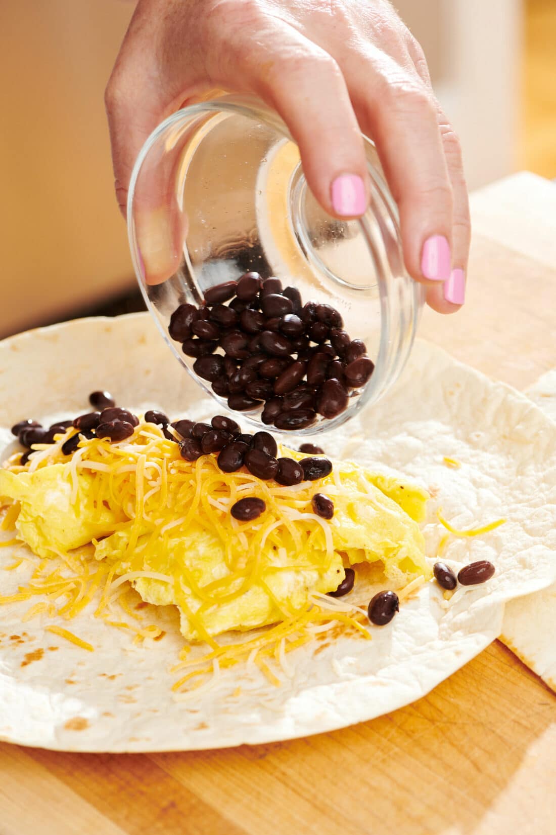 Woman pouring black beans onto a tortilla with egg and cheese.