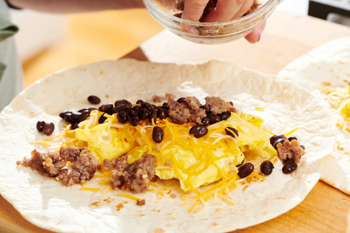 Sausage being put onto a tortilla with beans, eggs, and cheese.