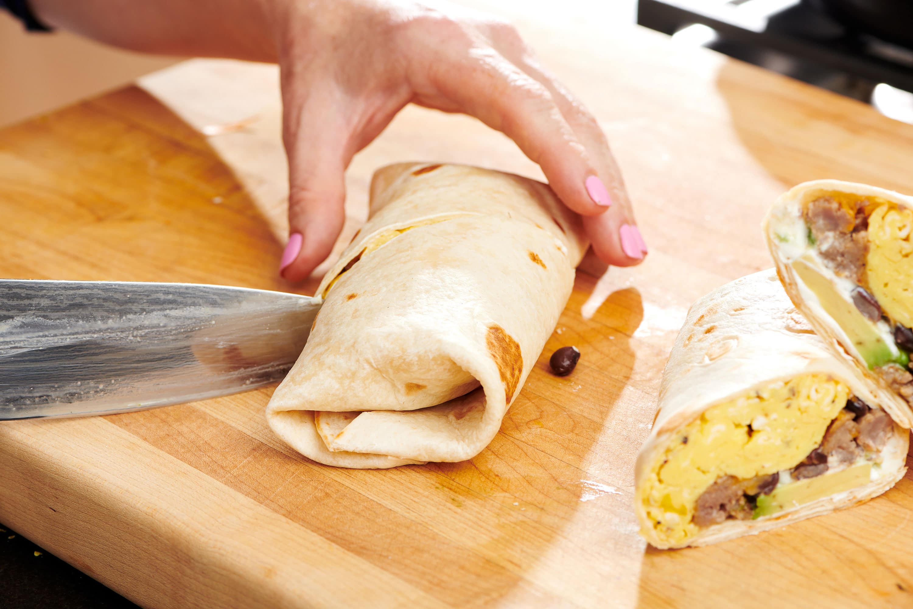 Woman slicing Breakfast Burrito with knife on cutting board.