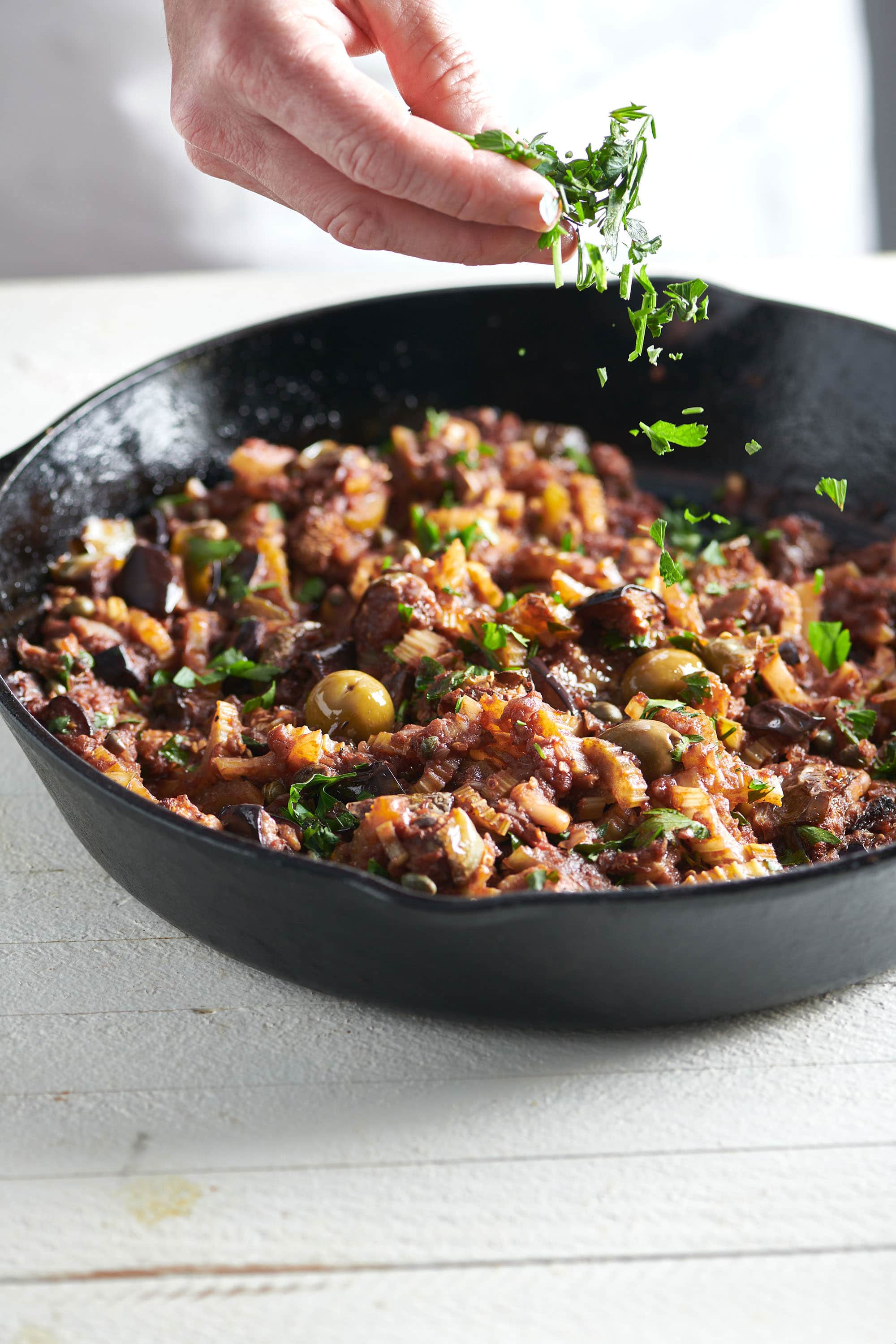 Caponata in a cast iron skillet being sprinkled with fresh parsley.