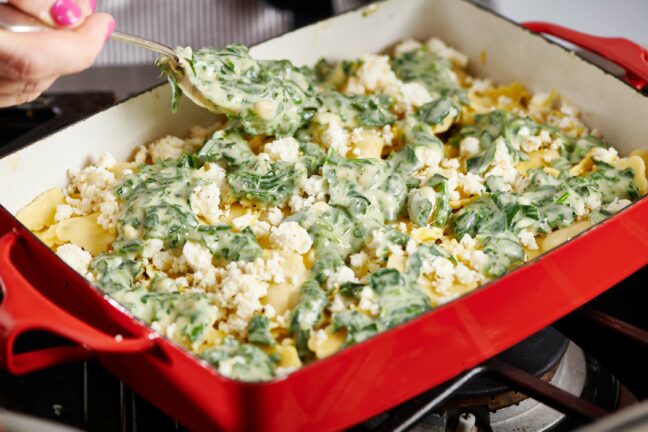 Spoon spreading creamy spinach mixture over a pan of ravioli.