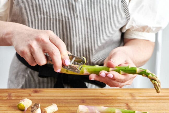 Woman using vegetable peeler to trim thick asparagus spears before cooking.