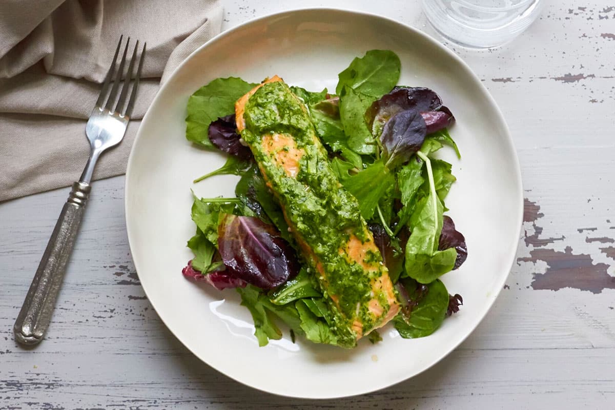 Salmon with herb sauce over salad on white plate.