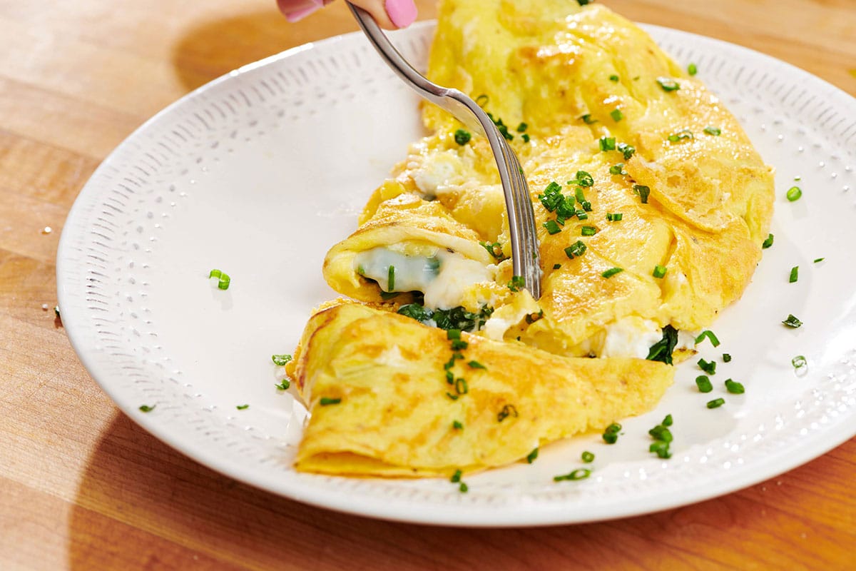 Fork cutting piece of a homemade omelet filled with feta and spinach with fresh chive garnish.