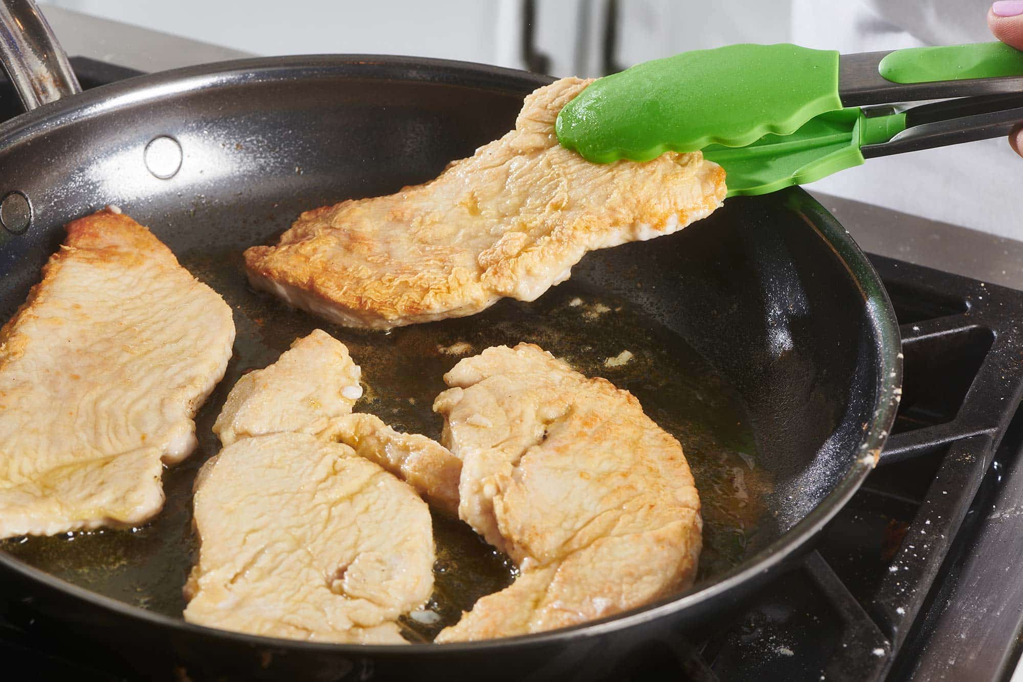 Green tongs flipping a turkey cutlet in a skillet.