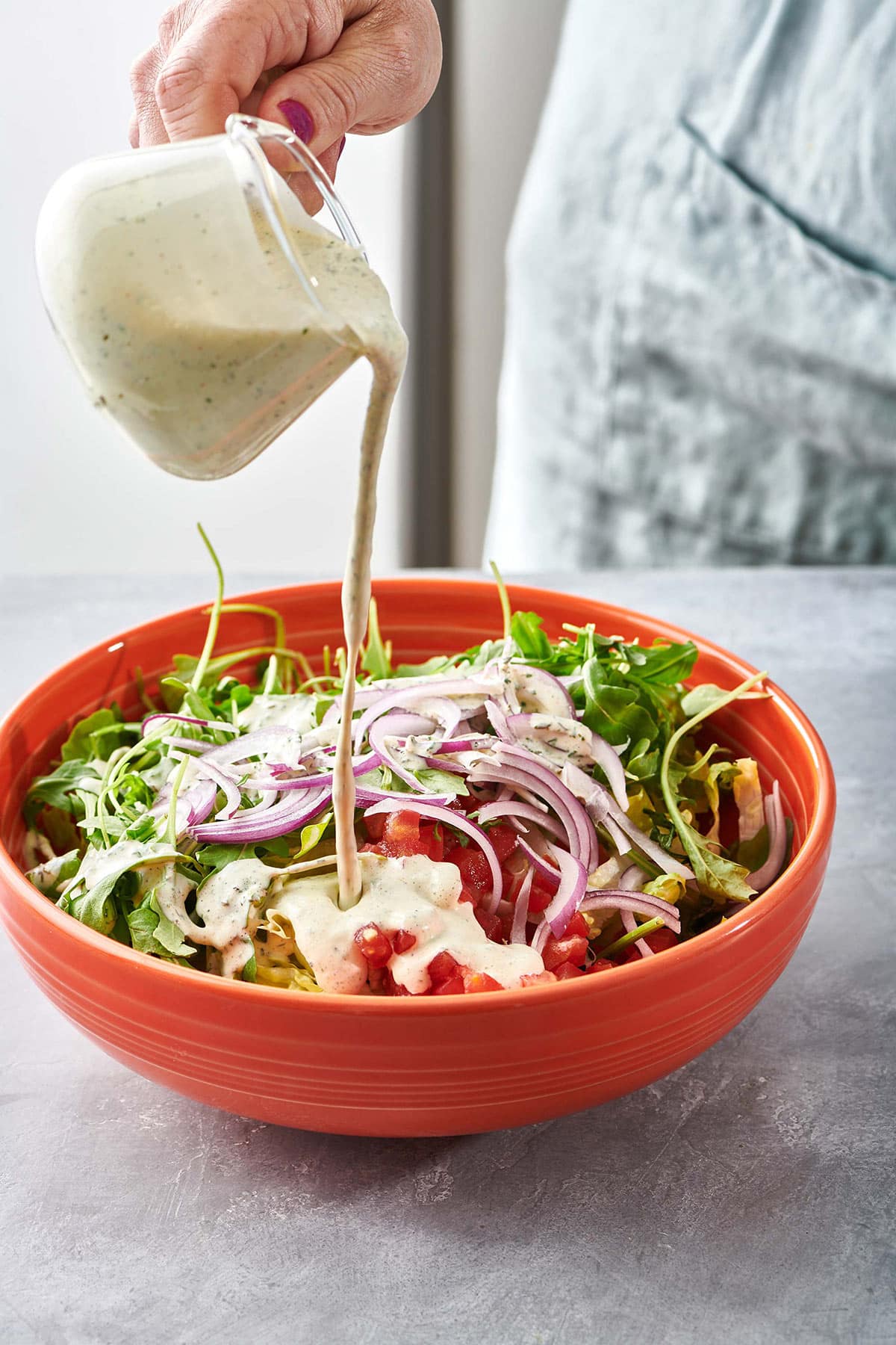 Woman pouring ranch dressing from small pitcher over salad.