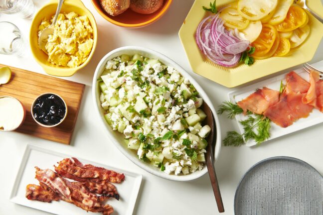 Bowl of Honeydew-Cucumber Salad with Feta on a table with bacon, bread, and other foods.