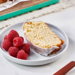 Slice of glazed lemon cake on plate with raspberries.
