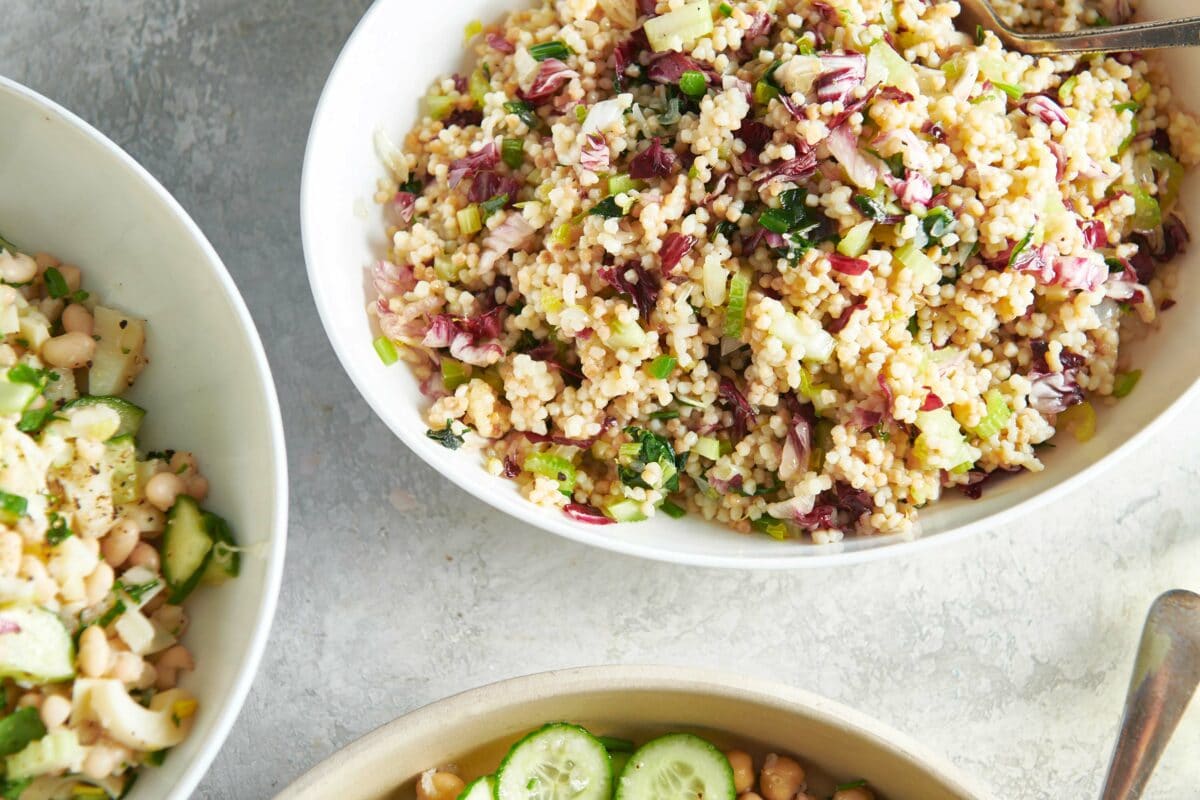 White bowl of Couscous Salad on a table with chickpeas and cucumbers.