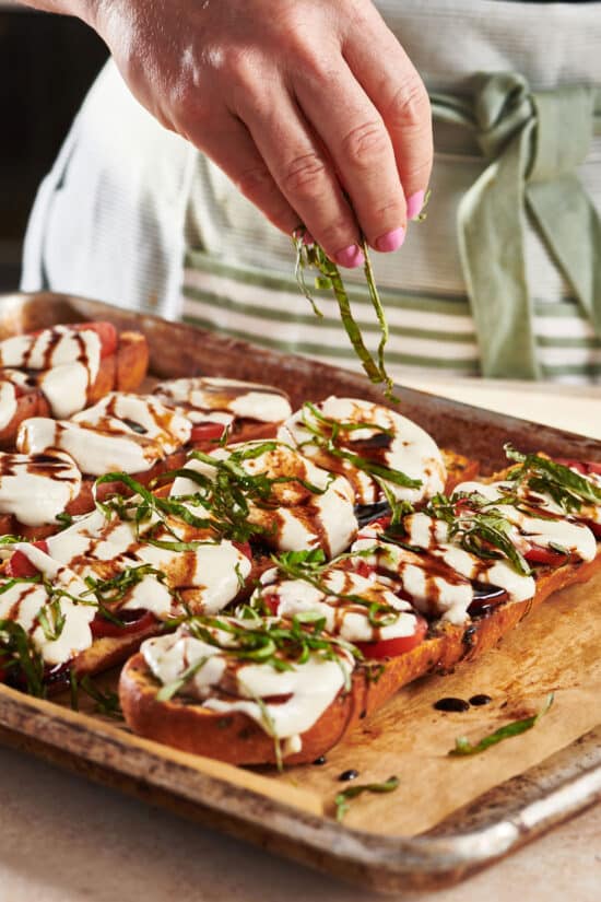 Woman sprinkling thinly sliced basil onto a tray of Caprese Garlic Bread.