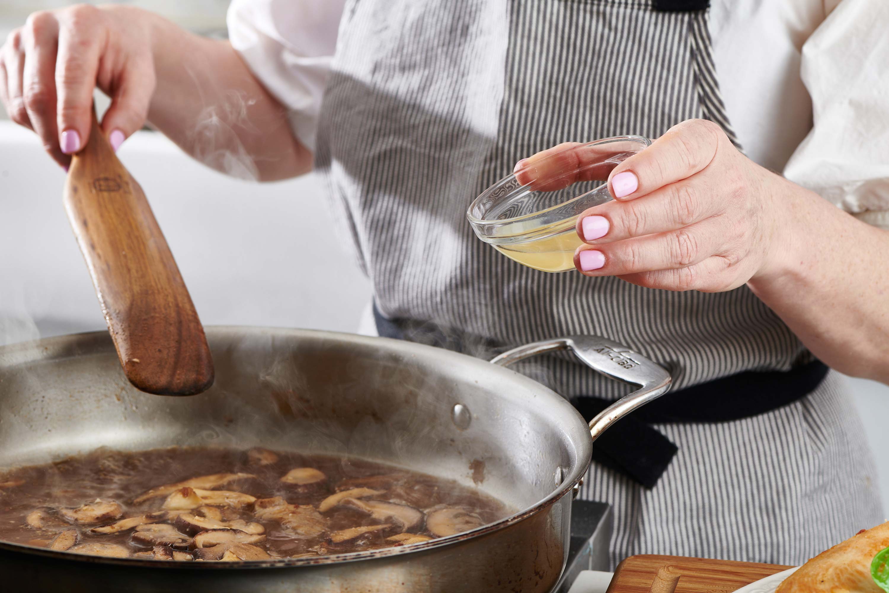 Woman holding a small bowl of lemon juice over a skillet of mushrooms.