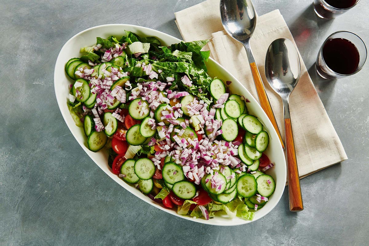 White, oblong bowl of Greek-inspired salad on table with drinks, silverware, and napkin.