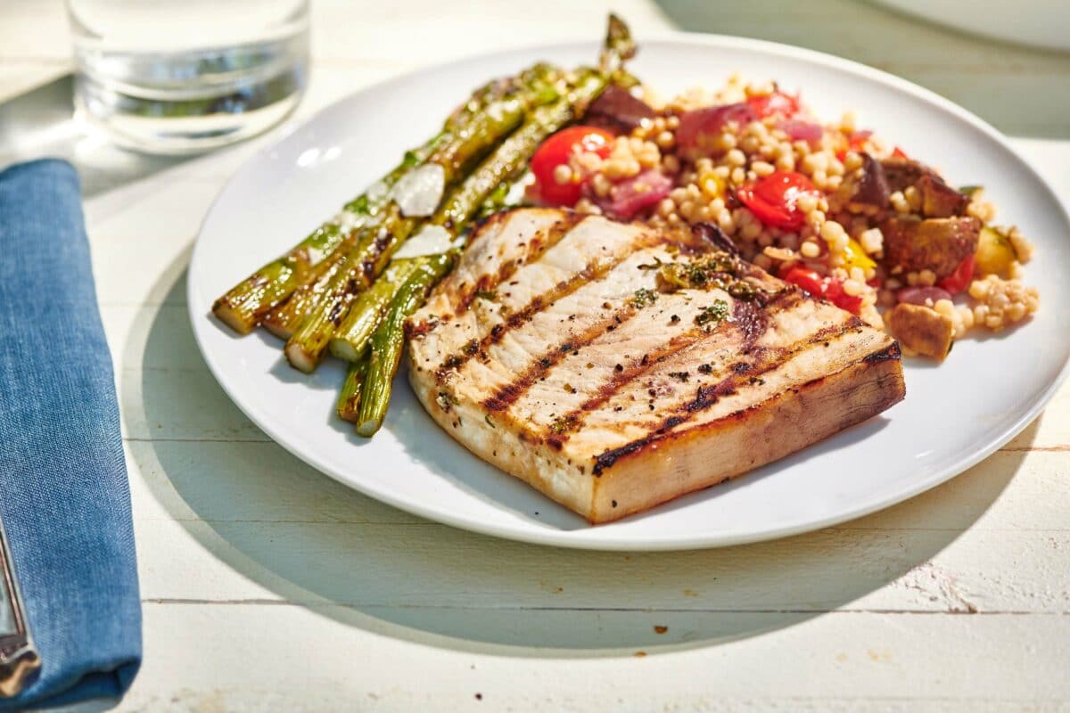 Plate of Grilled Swordfish and sides on a white, wooden table.