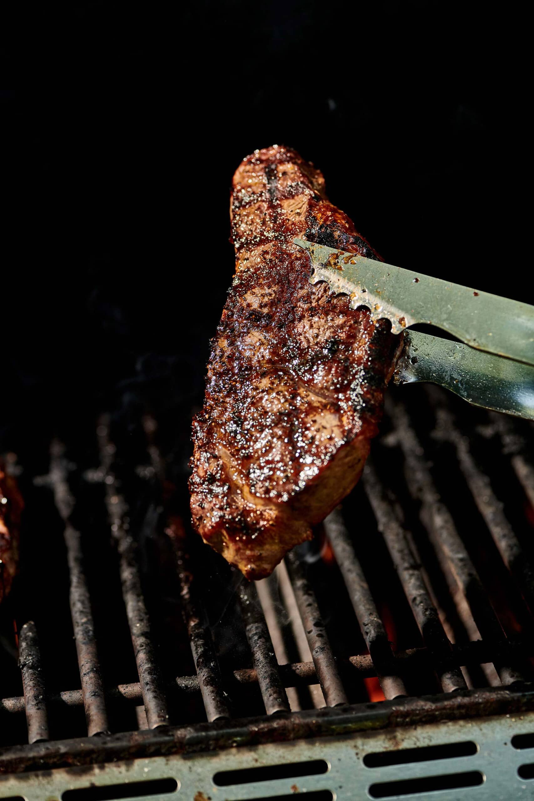 Tongs holding grilled New York strip steak over grill.