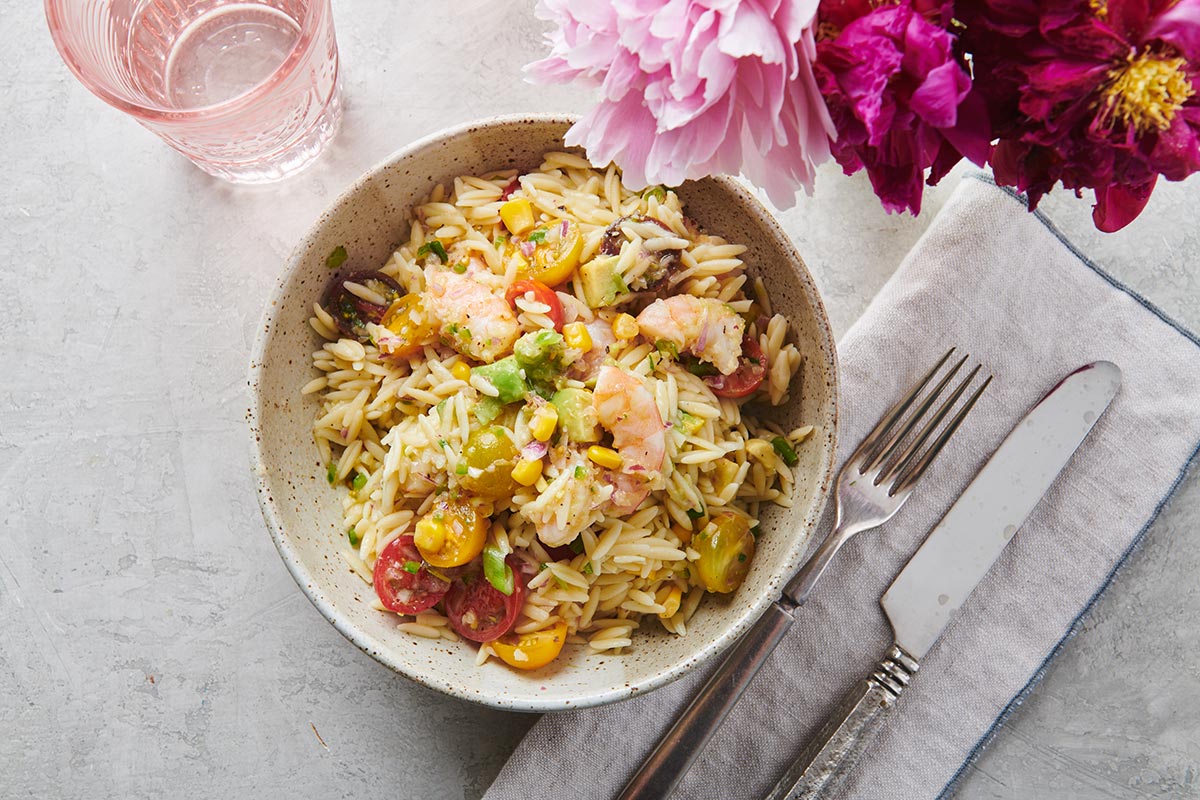 Bowl of shrimp orzo salad on table with silverware and pink flowers.