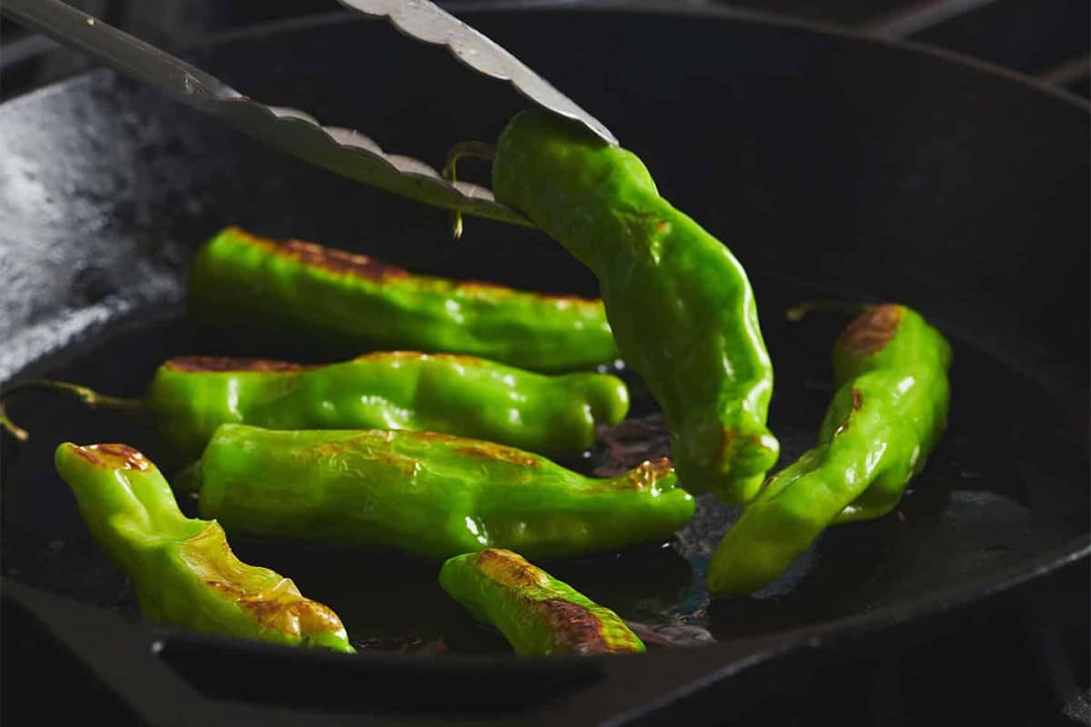 Tongs flipping a Shishito Pepper in a skillet.