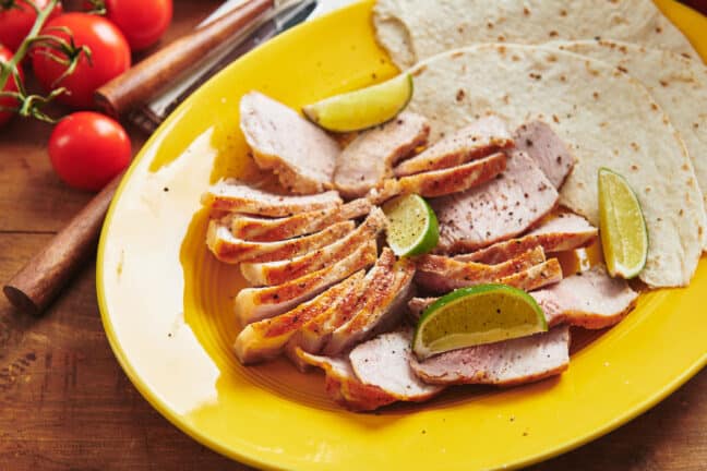 Grilled pork, lime wedges, and tortillas on a yellow plate.