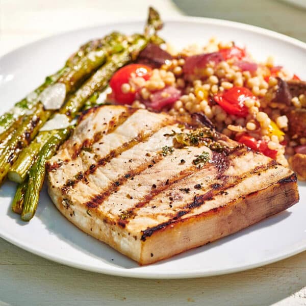 Plate of Grilled Swordfish and sides on a white, wooden table.