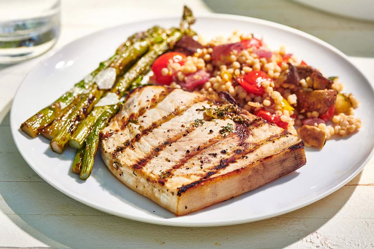 Plate of Grilled Swordfish and sides on a white, wooden table.