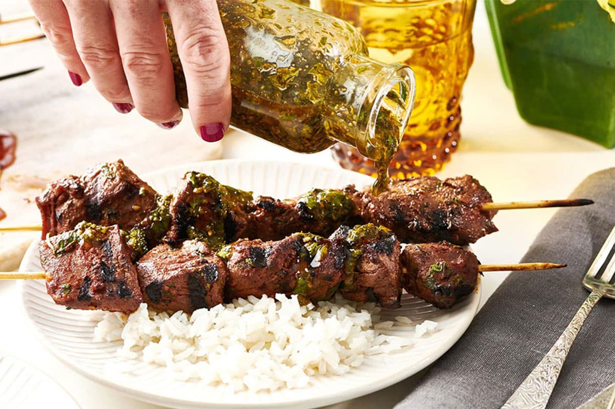 Woman pouring Chermoula sauce on Lamb Kebabs.