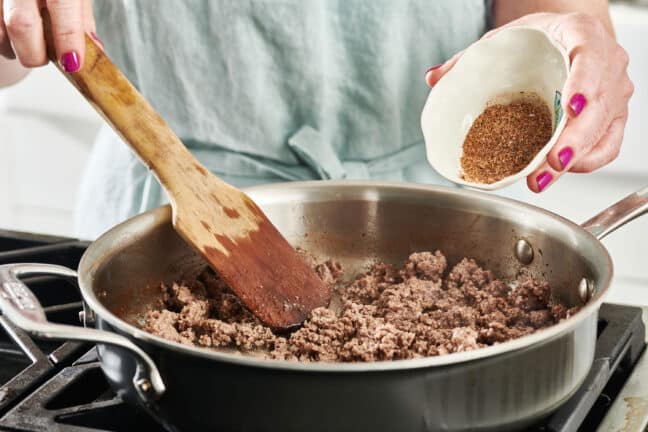 Woman adding spices to a skillet of ground beef.