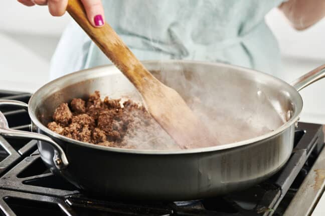 Wooden spatula stirring a steaming skillet of ground beef taco meat.