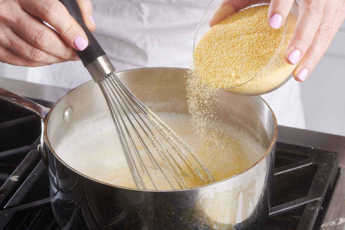 Woman pouring dry polenta into pot of milk and broth on stove.