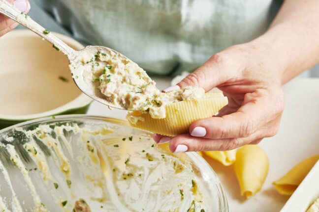 Woman spooning ricotta mixture into a pasta shell.