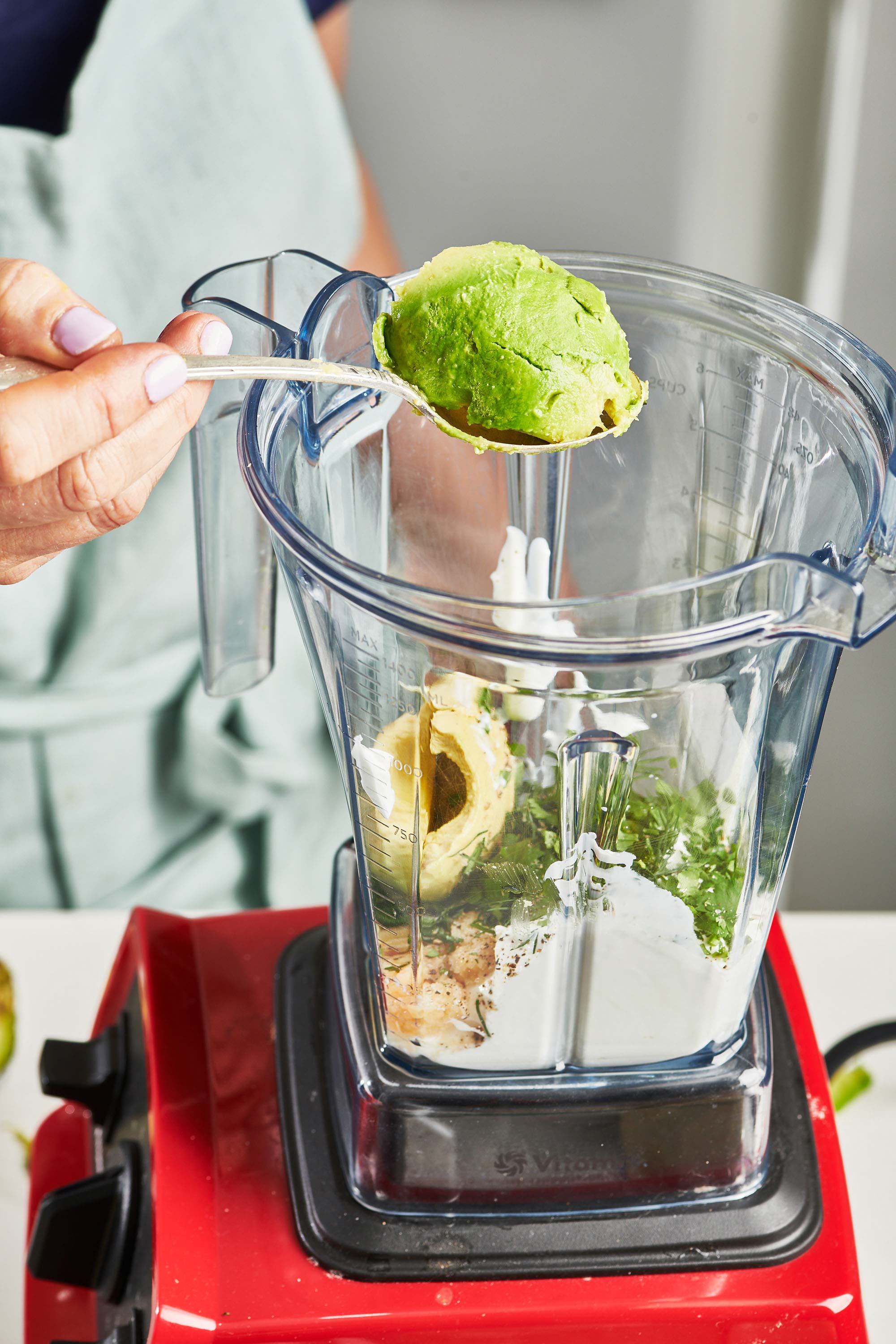 Woman holding avocado on a spoon over a blender.