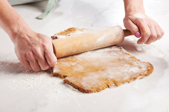 Woman using a wooden rolling pin on gingerbread dough.