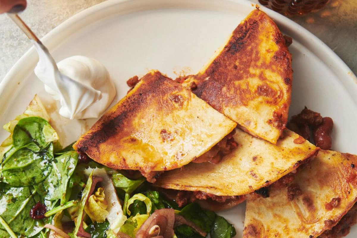 Spoon scooping sour cream onto a plate of Beefy Chili Cheese Quesadillas and salad.