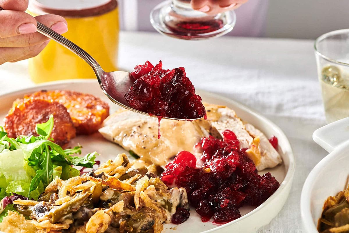 Spoon of Cranberry Sauce over a plate of Thanksgiving food.