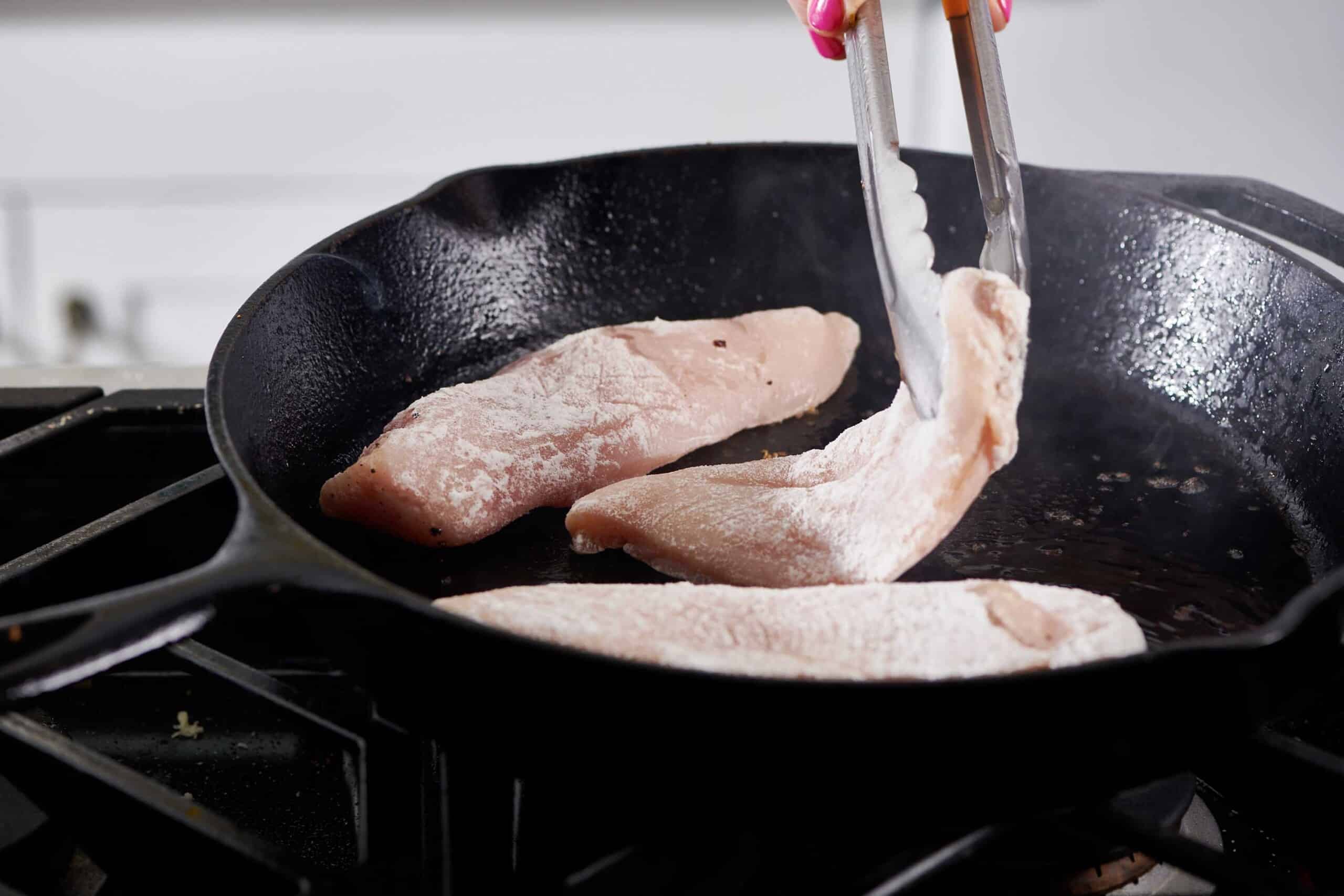 Tongs flipping chicken breast in a skillet.