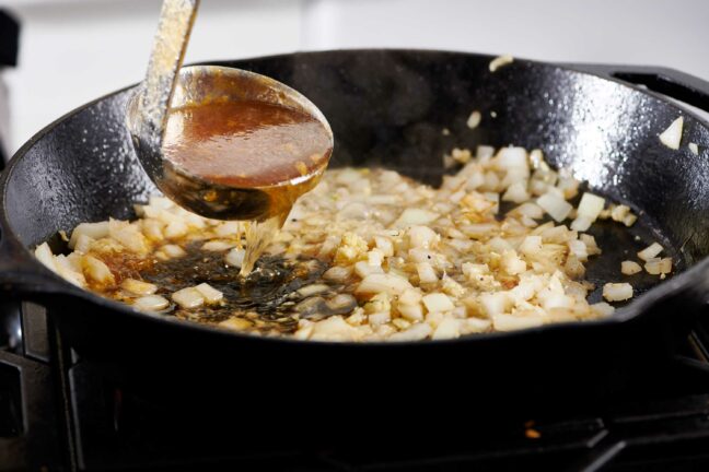 Ladle pouring broth into a skillet of diced onions.