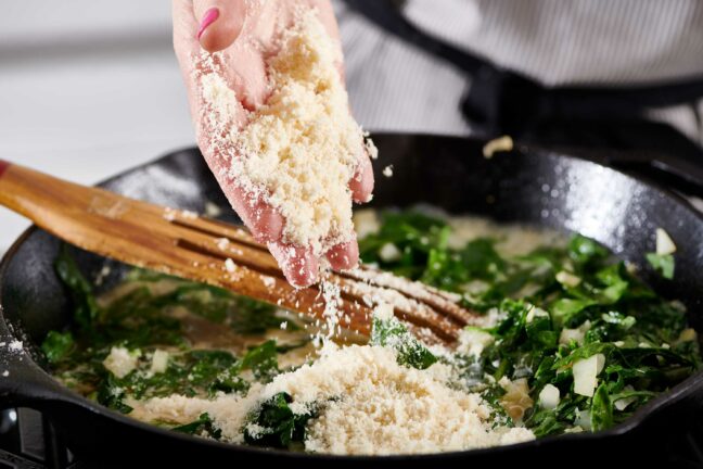 Woman tossing parmesan cheese into a skillet with spinach and onions.